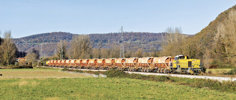 Partie du lieu de chargement situé à Gourdan- Polignan, la G 1206 708 achemine le train de ballast vers Bagiry où il permettra le ballastage de la voie posée. En arrière-plan, le V 211 182-584 assure la pousse du convoi. Le train passe à la hauteur de Bertren dans la vallée de la Garonne (27 novembre 2024 ; P. Mauduit