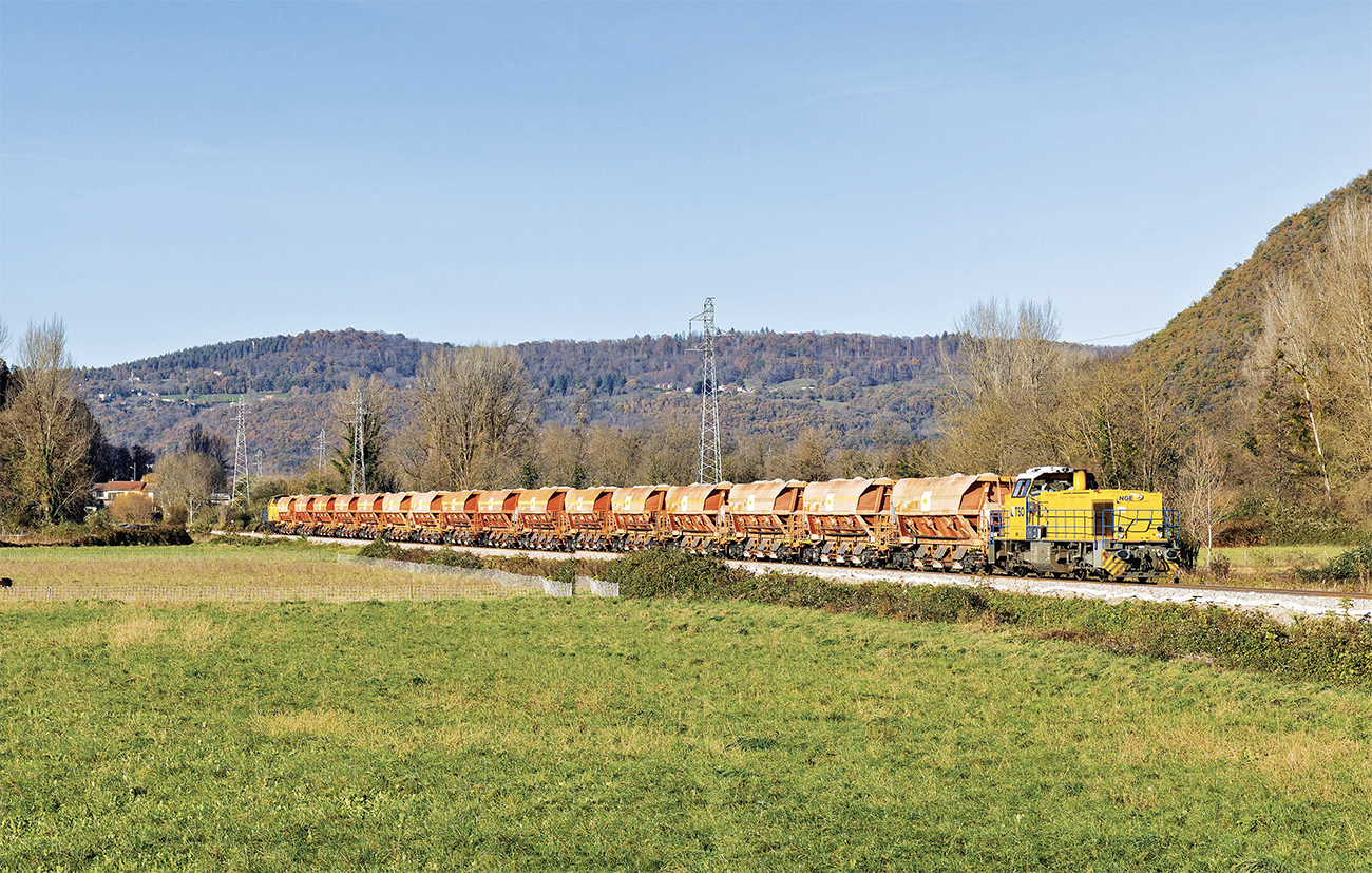 Partie du lieu de chargement situé à Gourdan- Polignan, la G 1206 708 achemine le train de ballast vers Bagiry où il permettra le ballastage de la voie posée. En arrière-plan, le V 211 182-584 assure la pousse du convoi. Le train passe à la hauteur de Bertren dans la vallée de la Garonne (27 novembre 2024 ; P. Mauduit