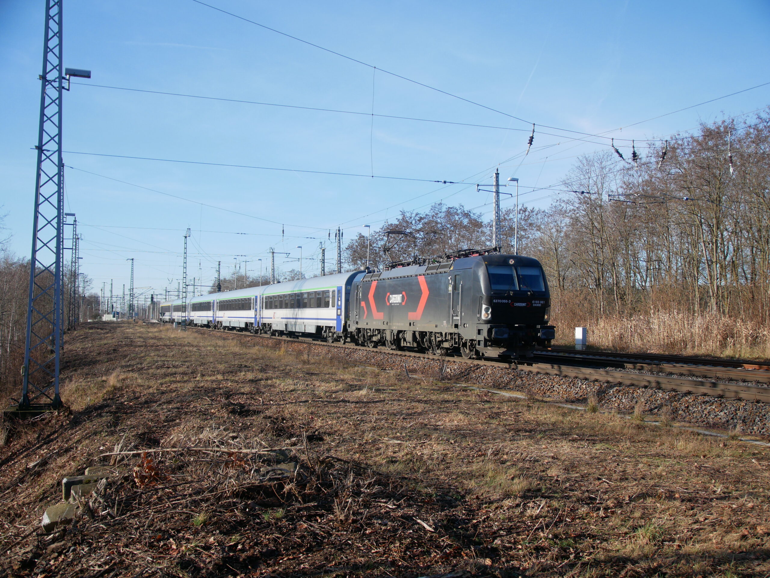 Le train EC 135 Leipzig Hbf – Wrocław Głowny, composé de la Vectron 5370 055-3 louée par PKP IC à la Rosco Cargounit remorquant une rame de voitures de PKP IC, vient de quitter à 11 h 55 la gare d’Elsterwerda (15 décembre 2025).