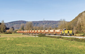 Partie du lieu de chargement situé à Gourdan- Polignan, la G 1206 708 achemine le train de ballast vers Bagiry où il permettra le ballastage de la voie posée. En arrière-plan, le V 211 182-584 assure la pousse du convoi. Le train passe à la hauteur de Bertren dans la vallée de la Garonne (27 novembre 2024 ; P. Mauduit